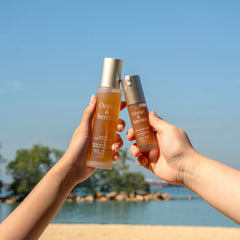 Two hands holding bottles of skincare products against a blue sky and ocean