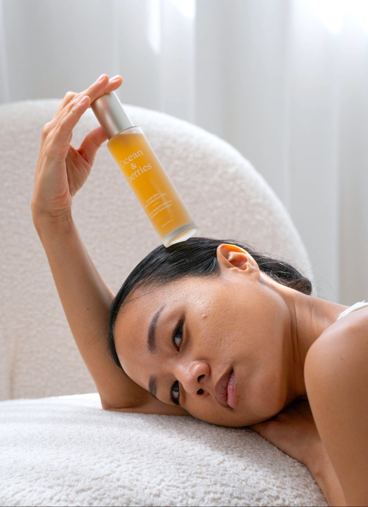 Woman holding a skincare bottle above her head with curtains in background